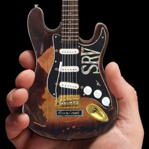 May include: A miniature replica of a vintage electric guitar with a distressed brown finish. The guitar has a black pickguard with a white "SRV" inscription and a gold "Custom" inscription on the body.