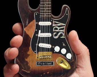 May include: A miniature replica of a vintage electric guitar with a distressed brown finish. The guitar has a black pickguard with a white "SRV" inscription and a gold "Custom" inscription on the body.