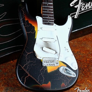May include: A black and orange electric guitar with a cracked paint finish. The guitar has a white pickguard and a Fender logo on the headstock. The guitar is made by Axe Heaven.
