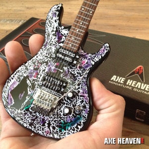 May include: A black electric guitar with a white and purple patterned design. The guitar has a black body and a maple neck. The guitar is a miniature replica of a real guitar. The guitar is made by Axe Heaven.