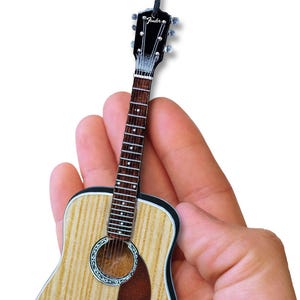 May include: A miniature acoustic guitar ornament with a wood grain finish, black headstock, and the word "Fender" printed on the headstock. The guitar is held in a hand.