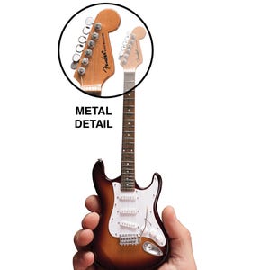 May include: A miniature replica of a Fender Stratocaster electric guitar, with a sunburst finish, white pickguard, and chrome hardware. The guitar is being held in a person's hand to show its size.