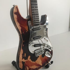 May include: A miniature electric guitar with a distressed, burnt orange finish. The guitar has a silver pickguard and chrome hardware. The guitar is on a black stand.