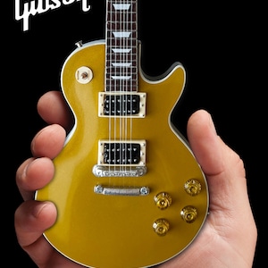 Puede incluir: Una guitarra eléctrica Gibson Les Paul dorada con herrajes cromados y una golpeador negro. La guitarra está siendo sostenida en la mano de una persona.