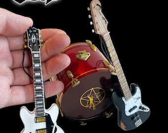 Op de afbeelding: Miniatuurreplica van een witte elektrische gitaar, een zwarte basgitaar en een rode basdrum met een gouden ster en een figuur in het midden. De drum heeft het woord "Rush" in rode letters aan de zijkant.