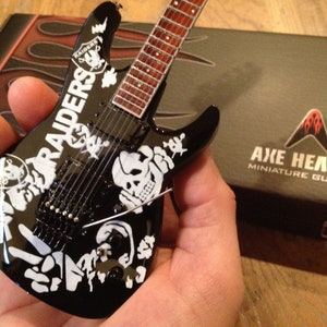 Könnte beinhalten: Schwarze elektrische Gitarre im Miniaturformat mit weißem Raiders-Logo und Totenkopf-Grafiken. Die Gitarre befindet sich in einer Hand und die Schachtel trägt den Schriftzug Axe Heaven Miniature Guitars.