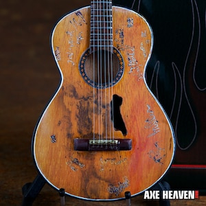 Pode incluir: Guitarra acústica com acabamento em madeira envelhecida, com assinaturas e boca preta. A guitarra tem uma borda branca e é exibida em um suporte preto. O logotipo Axe Heaven é visível no canto inferior direito.