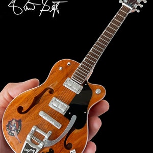 Puede incluir: Una guitarra el&eacute;ctrica marr&oacute;n y negra con una golpeador negro y un puente plateado. La guitarra tiene una pegatina de una mujer con un atuendo de pin-up con las palabras "Lucky Lady" y un gato negro con la palabra "Digsby" en el cuerpo. La guitarra tambi&eacute;n tiene dos dados blancos en el cuerpo.