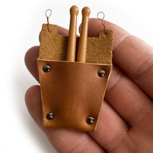 Puede incluir: Un porta baquetas de cuero marr&oacute;n con dos baquetas de madera en su interior. El porta baquetas tiene remaches met&aacute;licos y un lazo en la parte superior para colgar.