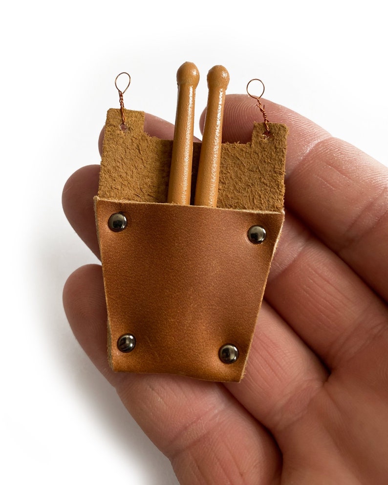 Puede incluir: Un porta baquetas de cuero marr&oacute;n con dos baquetas de madera en su interior. El porta baquetas tiene remaches met&aacute;licos y un lazo en la parte superior para colgar.