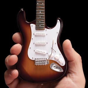 May include: A miniature brown and white electric guitar with a white pickguard. The guitar is being held in someone's hand.