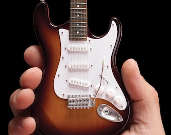 May include: A miniature brown and white electric guitar with a white pickguard. The guitar is being held in someone's hand.