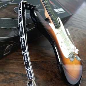 May include: A miniature Fender Stratocaster electric guitar with a sunburst finish and a black strap with the Fender logo.