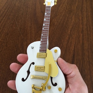 Peut inclure: Une guitare électrique blanche avec des accastillages dorés et une touche en bois brun. La guitare a un corps creux et une seule découpe.