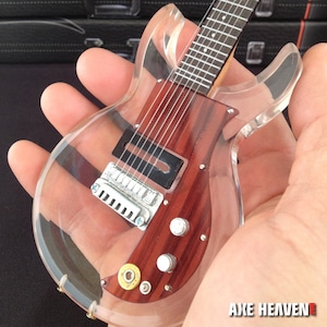 Könnte beinhalten: Eine transparente E-Gitarre aus Acryl mit einem Holzkörper und verchromten Beschlägen. Die Gitarre hat einen einzigen Tonabnehmer und zwei Knöpfe. Die Gitarre wird in einer Hand gehalten. Der Text "AXE HEAVEN" ist in der unteren rechten Ecke des Bildes sichtbar.