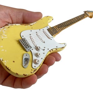 Könnte beinhalten: Eine Miniatur-E-Gitarre in Gelb mit einem Used-Look, weißem Schlagbrett und silberner Hardware. Die Gitarre hat drei Regler und einen Tremolo-Steg. Das Griffbrett ist braun, und die Kopfplatte ist hellgelb.