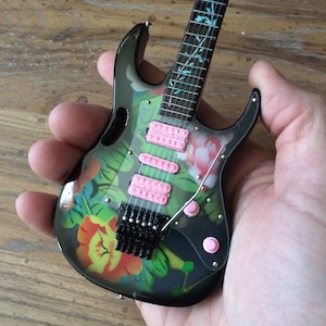 May include: A black electric guitar with a floral design and pink knobs. The guitar has a black body and a black headstock. The floral design is a mix of green, red, and yellow flowers.