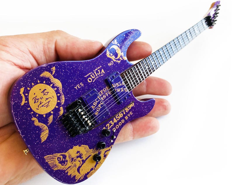 May include: A purple electric guitar with a gold Ouija board design. The guitar has a black pickguard and a gold sun and moon design on the body. The guitar is a miniature replica of a real guitar.