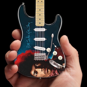May include: A miniature electric guitar with a blue and black finish featuring a portrait of Prince. The guitar has a white pickguard and a signature that reads "Both Sides of the Sky".
