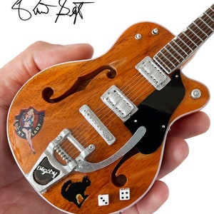 Puede incluir: Una guitarra el&eacute;ctrica en miniatura, de color marr&oacute;n, de cuerpo hueco, con una placa de protecci&oacute;n negra. La guitarra tiene un puente y clavijas de afinaci&oacute;n plateadas. La guitarra tiene varias pegatinas, incluyendo una pegatina de "Lucky Lady", una pegatina de "Big Sky" y una pegatina de un gato negro. La guitarra tambi&eacute;n tiene dos pegatinas de dados blancos.