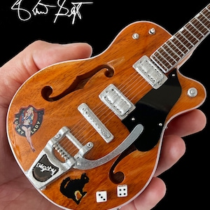 Puede incluir: Una guitarra el&eacute;ctrica en miniatura, de color marr&oacute;n, de cuerpo hueco, con una placa de protecci&oacute;n negra. La guitarra tiene un puente y clavijas de afinaci&oacute;n plateadas. La guitarra tiene varias pegatinas en el cuerpo, incluyendo una pegatina de una chica pin-up que dice "Lucky Lady", una pegatina de un gato negro y dos pegatinas de dados.