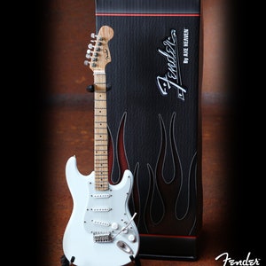Puede incluir: Una guitarra eléctrica Fender Stratocaster blanca con un mástil de arce y herrajes cromados. La guitarra está en un soporte y delante de una caja negra con el logotipo de Fender y el texto "By Axe Heaven".