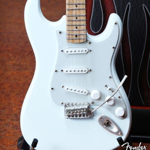 May include: A light blue electric guitar with a white pickguard and chrome hardware. The guitar has three white pickups and a tremolo bridge. The Fender logo and "Axe Heaven" text are visible on the lower right.