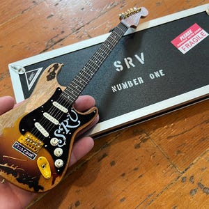 Op de afbeelding: Een miniatuurreplica van Stevie Ray Vaughan's iconische "Number One" gitaar. De gitaar is een Fender Stratocaster met een sunburst-afwerking en een zwarte pickguard. De gitaar heeft een witte "SRV"-handtekening op de body. De gitaar zit in een miniatuur gitaarkoffer met een "Fragile"-label.