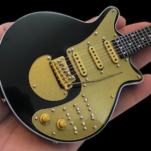 May include: A black electric guitar with gold hardware and a gold-colored body. The guitar has a unique shape and a single cutaway.