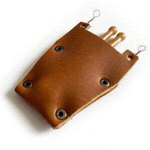 Puede incluir: Una funda de cuero marr&oacute;n con tres agujas de tejer de madera. La funda tiene remaches met&aacute;licos y lazos para colgar.