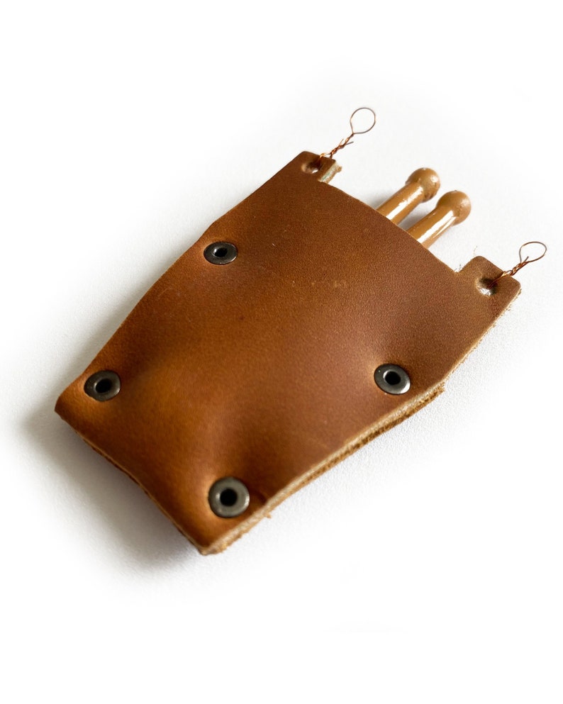 Puede incluir: Una funda de cuero marr&oacute;n con tres agujas de tejer de madera. La funda tiene remaches met&aacute;licos y lazos para colgar.