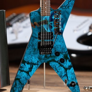 May include: A miniature electric guitar with a blue and black marbled design. The guitar features the text "PANTERA FAR BEYOND DRIVEN" in brown lettering. The guitar is displayed on a wooden surface.