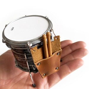 Puede incluir: Un set de bater&iacute;a en miniatura con una correa de cuero marr&oacute;n y dos baquetas de madera. El tambor es negro y blanco con un borde plateado.