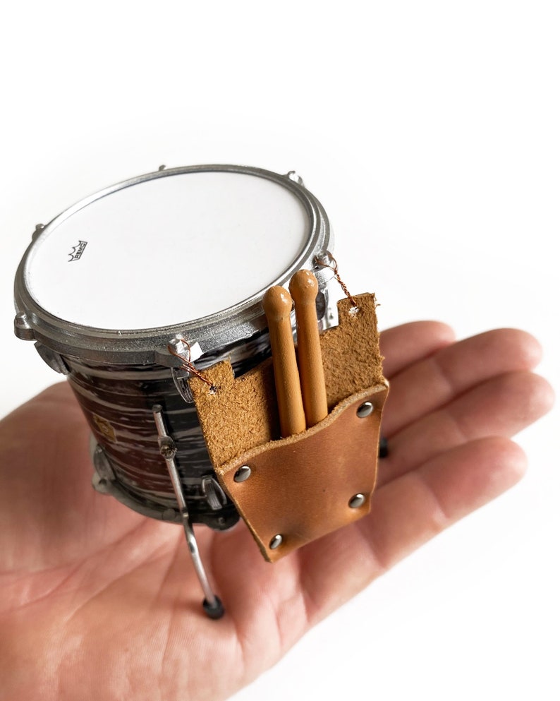 Puede incluir: Un set de bater&iacute;a en miniatura con una correa de cuero marr&oacute;n y dos baquetas de madera. El tambor es negro y blanco con un borde plateado.
