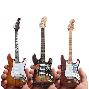 May include: Three miniature electric guitars, each with a different finish. The guitars are held in the hands of three people. The guitars are all Fender Stratocasters and have the name "SRV" written on the body.