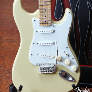 May include: A cream-colored electric guitar with a white pickguard and chrome hardware. The guitar has a maple neck and a rosewood fingerboard. The guitar is on a stand and has the text "Fender by Axe Heaven" on the bottom.