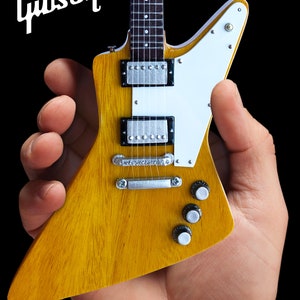 May include: A miniature Gibson Explorer electric guitar with a natural wood finish and white pickguard. The guitar is held in a hand.