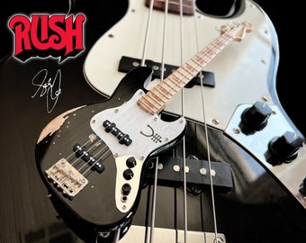 Op de afbeelding: Een zwarte en witte elektrische basgitaar met een versleten afwerking. De gitaar heeft een witte pickguard met een handtekening en een symbool erop. De gitaar ligt op een grotere zwarte en witte basgitaar.