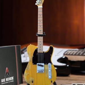 May include: A miniature yellow electric guitar with a black pickguard and a Fender logo on the headstock. The guitar is on a stand and has a worn, vintage look. The guitar is labeled "Axe Heaven Miniature Guitars".