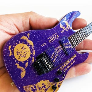 May include: A purple electric guitar with a gold Ouija board design. The guitar has a sun and moon design on the body and the words "YES" and "NO" on the headstock.