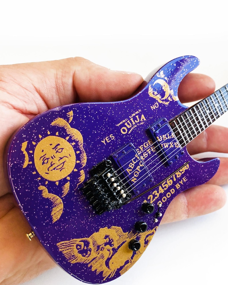 May include: A purple electric guitar with a gold Ouija board design. The guitar has a sun and moon design on the body and the words "YES" and "NO" on the headstock.