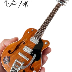 Puede incluir: Una guitarra el&eacute;ctrica en miniatura de color marr&oacute;n y negro con una placa de protecci&oacute;n negra y herrajes plateados. La guitarra tiene una pegatina de una chica pin-up con el texto "Lucky Lady" y un gato negro con el texto "Digsby".