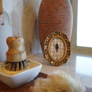 May include: An oval, gold-colored picture frame with a decorative border holds a ring and a decorative key. A large, brown vase stands behind the frame. A white soap dish with a wooden brush and a natural sponge are in the foreground.