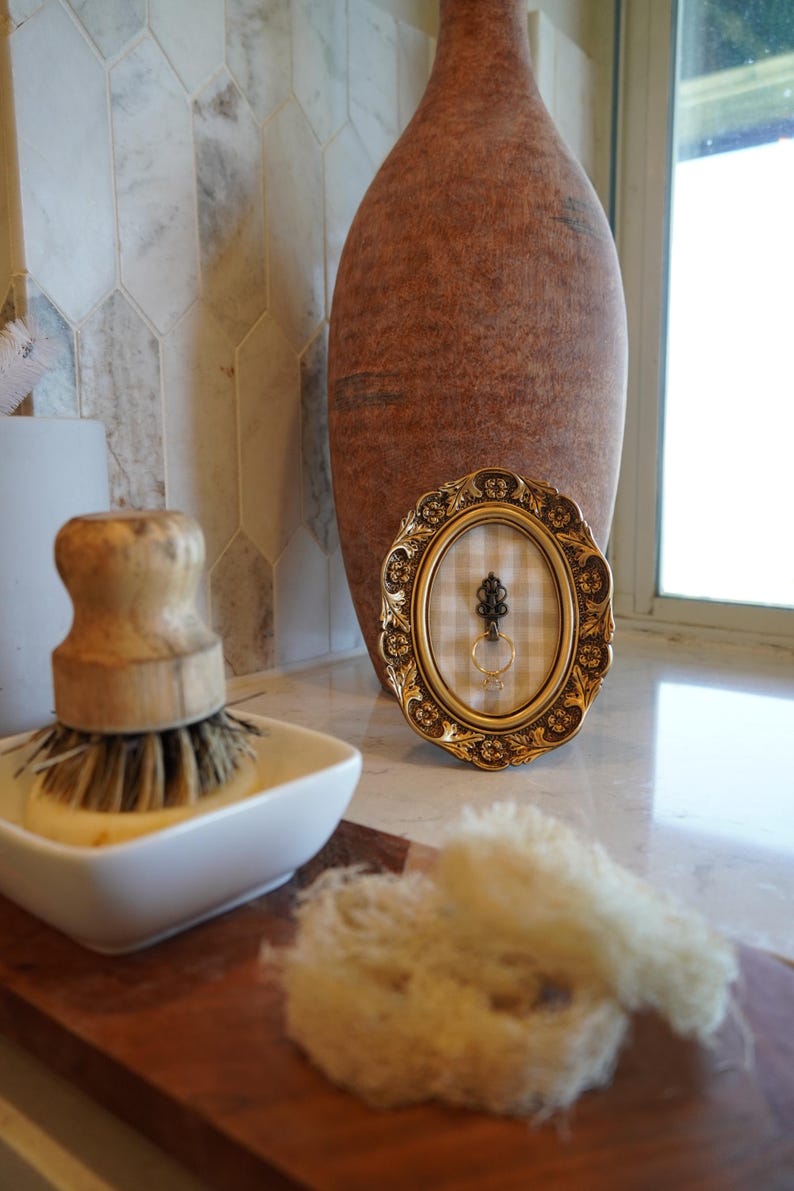 May include: An oval, gold-colored picture frame with a decorative border holds a ring and a decorative key. A large, brown vase stands behind the frame. A white soap dish with a wooden brush and a natural sponge are in the foreground.
