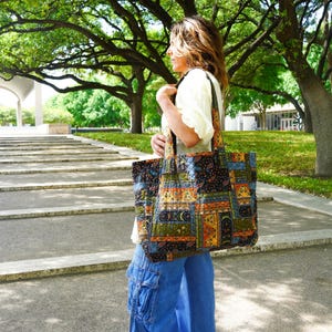 May include: A large, quilted tote bag with a patchwork design in shades of blue, green, orange, and black. The bag has a rectangular shape with a wide strap. The person is wearing blue jeans and a white shirt.