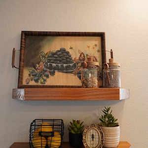 May include: Wooden shelves with decorative items. The top shelf holds a framed painting of blackberries, glass jars, and a wooden tray. The bottom shelf features a wire basket of lemons, a small succulent, a framed oval picture, and a potted plant.