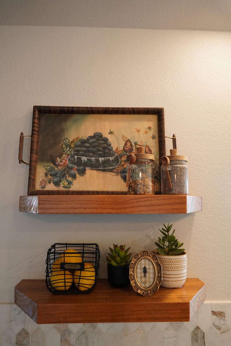May include: Wooden shelves with decorative items. The top shelf holds a framed painting of blackberries, glass jars, and a wooden tray. The bottom shelf features a wire basket of lemons, a small succulent, a framed oval picture, and a potted plant.