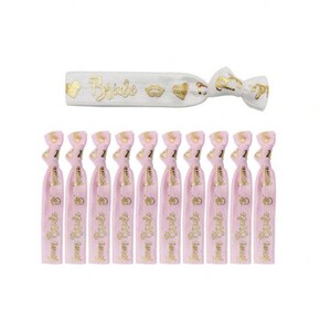 May include: Ten pink elastic hair ties with gold lettering that says "Bride Tribe" and a gold ring design. One white elastic hair tie with gold lettering that says "Bride" and gold crown and heart designs.