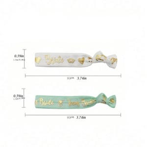 May include: Two hair ties with gold lettering. The first hair tie is white with the word "Bride" and gold hearts and lips. The second hair tie is light green with the words "Bride" and "Team Bride". Both hair ties are 3.74 inches long.