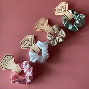 May include: Four satin scrunchies in different colors, each attached to a card with the text "To have and to hold your hair back". The cards are shaped like diamonds.